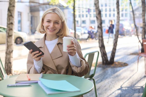 Portrét blond usměvavé ženy sedí v kavárně, těší šálek kávy, pomocí mobilního telefonu, pracuje na smartphone, sociální média blogger dělat příspěvek on-line. - Fotografie, Obrázek