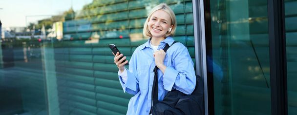 Életmód portré mosolygós fiatal női modell, diák hátizsákkal, vár valakit az utcán, áll a szabadban okostelefonnal. - Fotó, kép