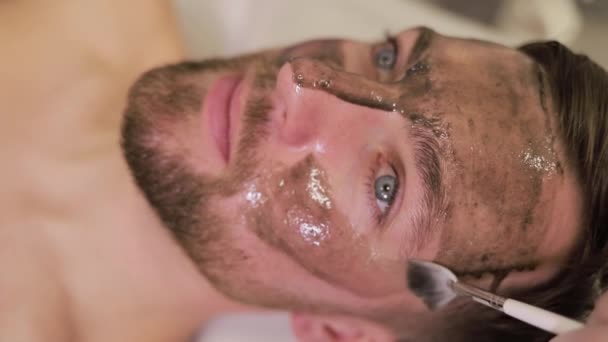 Mladý muž podstupuje masážní ošetření v kosmetických lázních. Neoholený muž s kosmetickou maskou péče v lázeňském salonu, boční, horní pohled. - Záběry, video