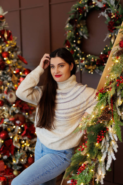 Retrato de mulher em jeans e suéter posando em quarto decorado de Natal escuro - Foto, Imagem