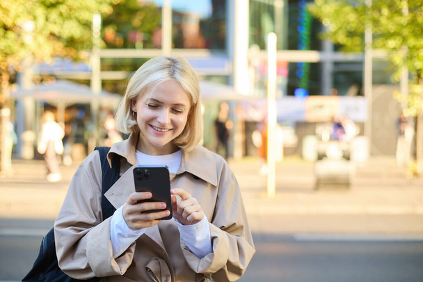 Nahaufnahme Porträt einer jungen lächelnden blonden Frau, Studentin auf der Straße, mit dem Handy, Nachrichten auf dem Smartphone checken, auf der Straße gehen. - Foto, Bild