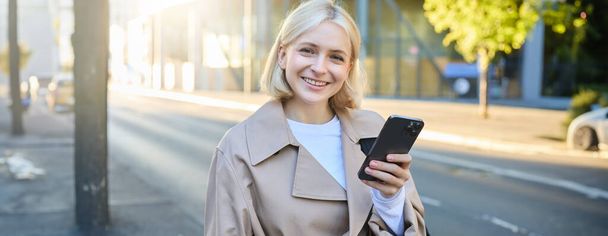 Portrait de femme blonde souriante, portant un trench coat dans la rue, regardant heureux, utilisant le téléphone portable, tenant smartphone, a l'expression du visage heureux et satisfait. - Photo, image