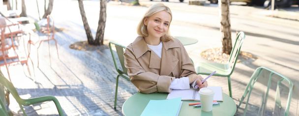 Image d'une jeune femme blonde dans une tranchée élégante, assise dans un café extérieur, buvant du café et travaillant sur des documents, étudiant, faisant ses devoirs dans son carnet, souriant à la caméra. - Photo, image