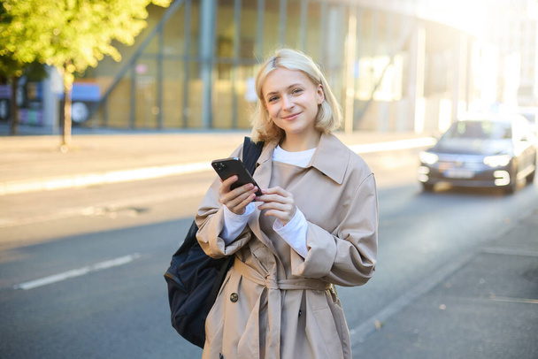 Retrato de estilo de vida de una mujer joven en la calle, de pie cerca de la carretera con coches, lleva mochila y sostiene el teléfono móvil, pide un paseo en la aplicación del teléfono inteligente, sonriendo y mirando feliz a la cámara. - Foto, imagen