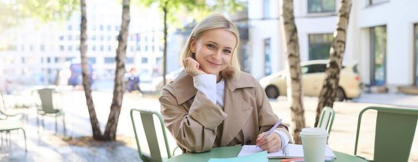 Portret młodej nowoczesnej kobiety w stylowym płaszczu, siedzącej w słoneczny dzień w kawiarni na świeżym powietrzu, piszącej w swoim notesie, pracującej lub odrabiającej lekcje, pijącej kawę. - Zdjęcie, obraz