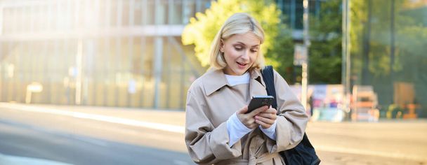 Retrato de menina surpresa na rua, mulher olhando para o telefone com sobrancelhas levantadas, lendo uma mensagem no smartphone, esperando por alguém perto da estrada, segurando mochila. - Foto, Imagem