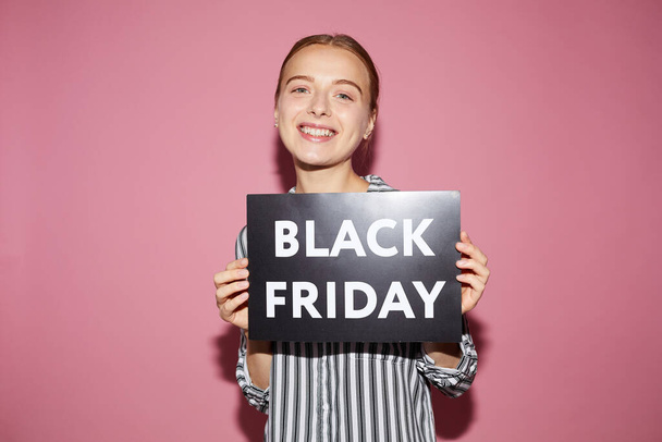 Cheerful beautiful young woman in stripped shirt standing against pink background and showing banner while welcoming to Black Friday sale - Zdjęcie, obraz