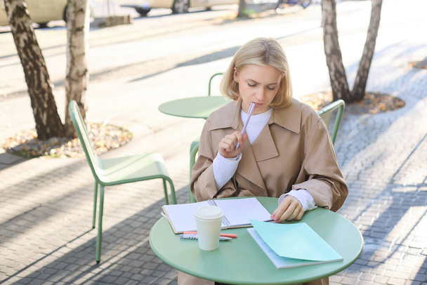 Ritratto di giovane donna che pensa mentre fa i compiti, seduta in un caffè all'aperto da sola, che beve caffè e scrive nel taccuino, che sembra premurosa, meditando smth. - Foto, immagini