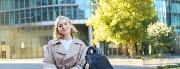 Vertikale Aufnahme einer jungen blonden Frau, die mit Handy auf einer Bank sitzt, Rucksack hat, auf jemanden wartet, Taxi in Smartphone-Anwendung bestellt und sich umschaut, unbeschwert lächelnd. - Foto, Bild