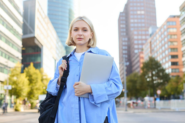 Kendine güvenen genç bir kadının portresi, sırt çantası ve dizüstü bilgisayarı olan üniversite öğrencisi, derse gidiyor, arkasında büyük binalar olan boş bir sokakta dikiliyor.. - Fotoğraf, Görsel