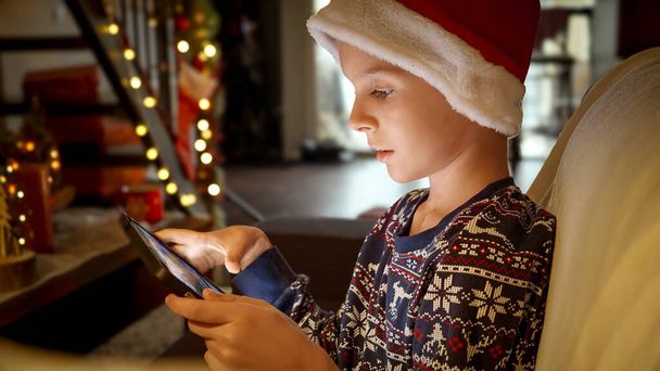 Close-up portret van kleine jongen in Santa 's hoed zitten op de bank en het spelen van games op tablet computer tijdens Kerstmis. - Foto, afbeelding