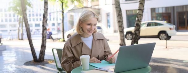 Mujer joven, freelancer, estudiante haciendo la tarea en la cafetería al aire libre, bebiendo su café en la calle, usando el ordenador portátil, se conecta a la conferencia en línea o haciendo curso en Internet. - Foto, Imagen