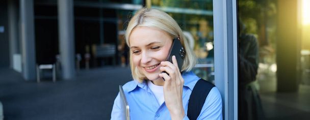 gelukkig vrouw model, blonde vrouw praten op mobiele telefoon, chatten met iemand, in de buurt van kantoor gebouw met laptop en smartphone, glimlachen. - Foto, afbeelding