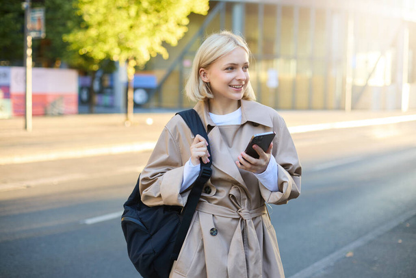 Modern young woman with backpack, student using smartphone, standing on street, sunny day, waiting for ride, using mobile phone application. - Photo, Image