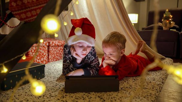 Dos niños pequeños viendo dibujos animados en la tableta de la noche de Navidad en la casa decorada con luces y guirnaldas. Vacaciones de invierno, celebraciones y fiestas - Foto, Imagen
