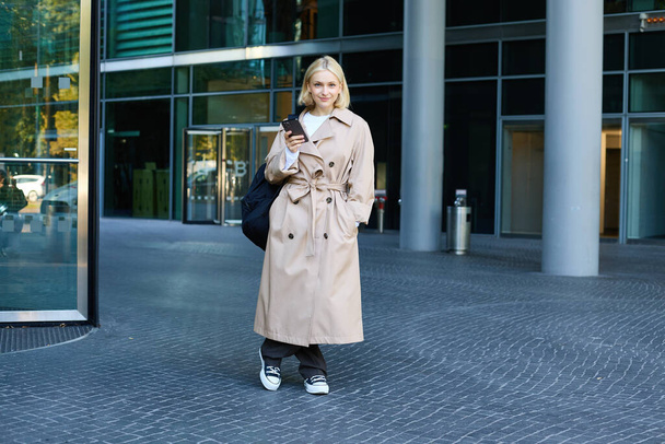 Beeld van stijlvolle, moderne jonge vrouw op straat, poseren in trench jas met rugzak, staande en lachende, holding smartphone. - Foto, afbeelding