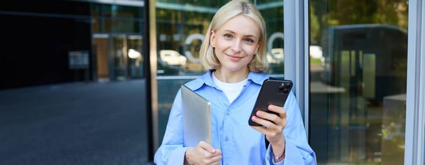 Portret van jonge vrouwelijke student die buiten de universiteitscampus staat, poseert in de buurt van gebouw, laptop en smartphone vasthoudt, blauw hemd draagt. - Foto, afbeelding