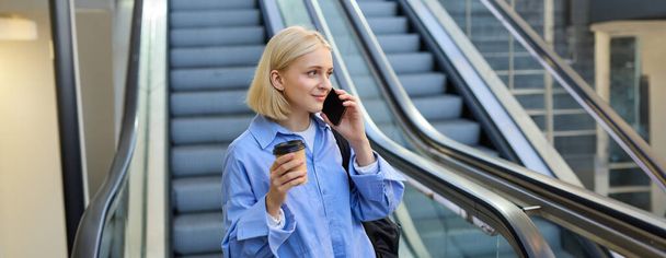 Lifestyle shot van jonge vrouwelijke student, vrouw praten op mobiele telefoon, koffie drinken, in de buurt van roltrap in het centrum van de stad. - Foto, afbeelding