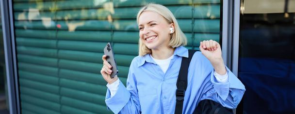 Женщина с рюкзаком и мобильным телефоном, в беспроводных наушниках, слушая музыку и улыбаясь, стоя на улице в центре города, смеясь. - Фото, изображение