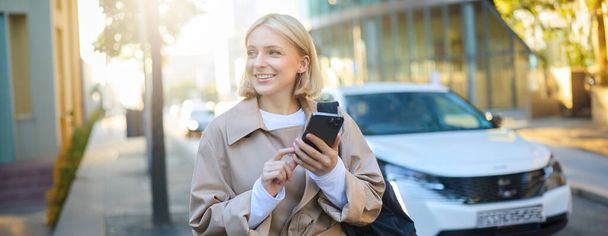 Portrait of young woman using map application on smartphone, student with backpack, walks along street on sunny day, holds mobile phone in hand. - Photo, Image