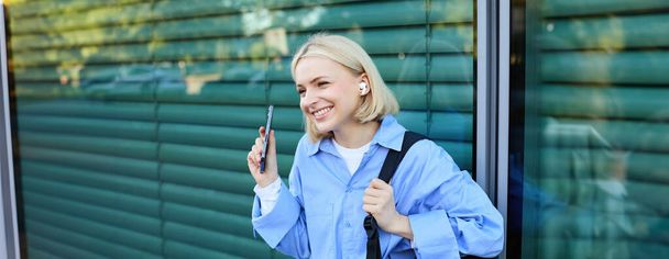 Primer plano retrato de modelo femenina rubia sonriente, estudiante universitaria en auriculares, escuchando música en la calle, sosteniendo el teléfono inteligente, esperando a alguien al aire libre. - Foto, imagen