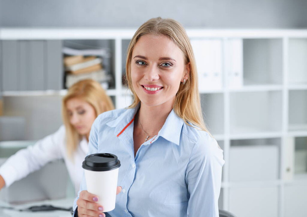 Mujer de negocios sonriente bebiendo café de una taza de papel en el retrato de la oficina mirando a la cámara. Sosteniendo en su mano y descansando durante un descanso. - Foto, imagen
