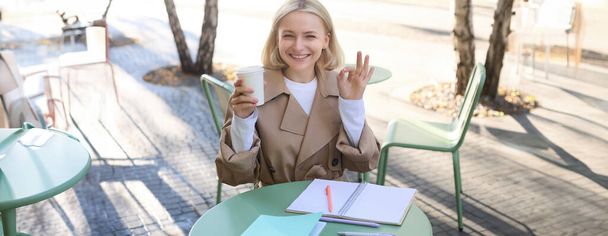 Fröhliche junge Studentin, Frau sitzt im Straßencafé, hält Tasse Kaffee, zeigt Okay-Zeichen, ok-Geste, macht Hausaufgaben, schreibt in Notizbuch. - Foto, Bild