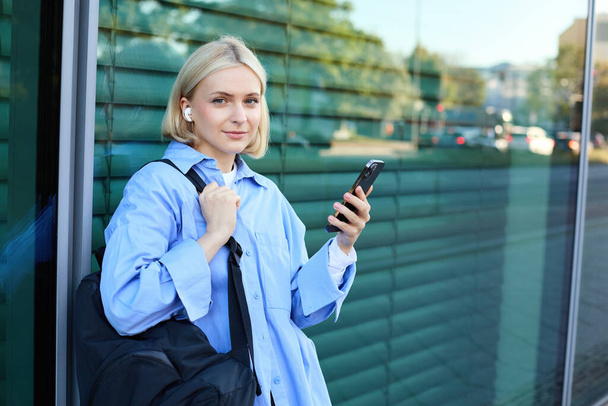 Stile di vita ritratto di giovane modella sorridente, studentessa con zaino, in attesa di qualcuno per strada, in piedi all'aperto con smartphone. - Foto, immagini