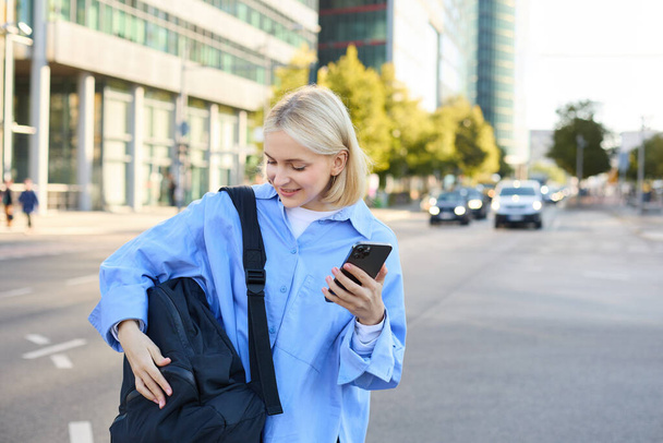 Életmód felvétel fiatal nőről, diák áll okostelefonnal az utcán, kinyitja a hátizsákját. Emberek és foglalkozás fogalma - Fotó, kép