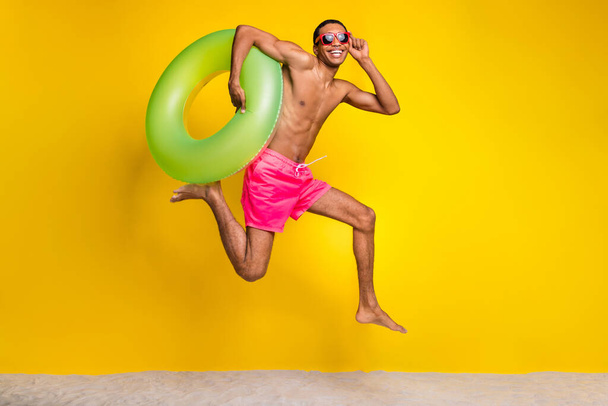 Full length photo of cheerful cool shirtless man jumping high holding inflatable circle isolated yellow color background. - Photo, image