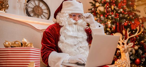 Papá Noel sentado en casa y leyendo el correo electrónico en el ordenador portátil con la lista de deseos de Año Nuevo - Foto, Imagen