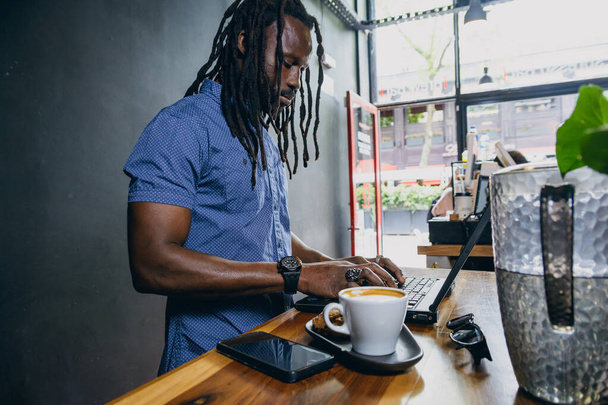 Hombre africano con rastas, barba y camisa, hombre de negocios autónomo, de pie en la cafetería trabajando en su computadora portátil y tomando café, espacio para copiar. - Foto, imagen