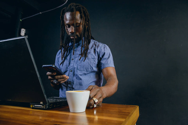 Hombre negro africano con rastas y barba, autónomo, de pie dentro de la cafetería utilizando el teléfono de trabajo y la celebración de la taza de café, la gestión de su negocio en línea, sirviendo a los clientes a través de Internet. - Foto, Imagen