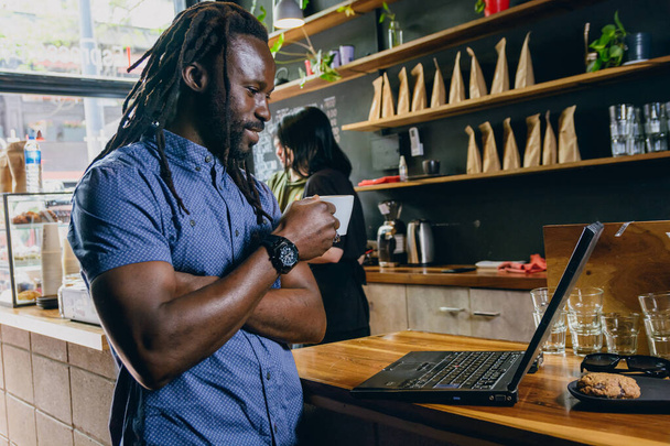 чорний бізнесмен з дредлоками і бородою, самозайнятий, щасливий стоїть на ноутбуці на лічильнику ресторану, сміється і тримає чашку кави в руках, простір для чашок, концепція способу життя - Фото, зображення