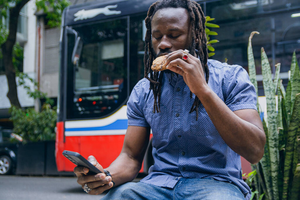 mladý černý Afričan s dredy a plnovousem, sedí s mobilním telefonem a kontroluje telefon a jí sušenky venku. koncept životního stylu, kopírovací prostor - Fotografie, Obrázek