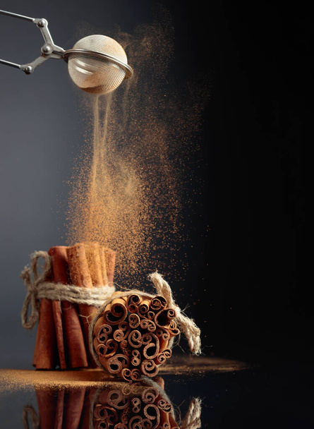 Cinnamon powder is poured out of the strainer. The ground cinnamon, cinnamon sticks, tied with jute rope on a black reflective background. Copy space. - Fotografie, Obrázek