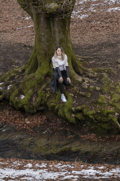 Karlı bir ormanda ağaç kökleri üzerinde oturan bir kadın. - Fotoğraf, Görsel