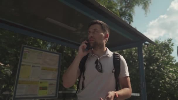 Man controleert de tijd op polshorloge in afwachting van de bus bij de halte, oproepen om te waarschuwen voor vertraagde afspraak, man communiceert via mobiele telefoon tijdens het vertragen van het vervoer in Duitsland. Een geïrriteerde man is laat..  - Video