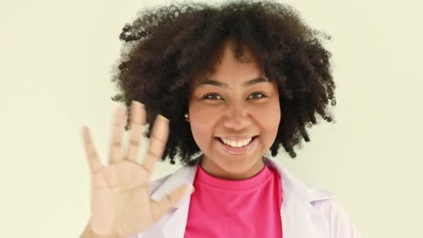 Cute African American female nutritionist sitting on a desk, smiling and raising her hand in greeting, sitting on a desk in a hospital office. - Footage, Video