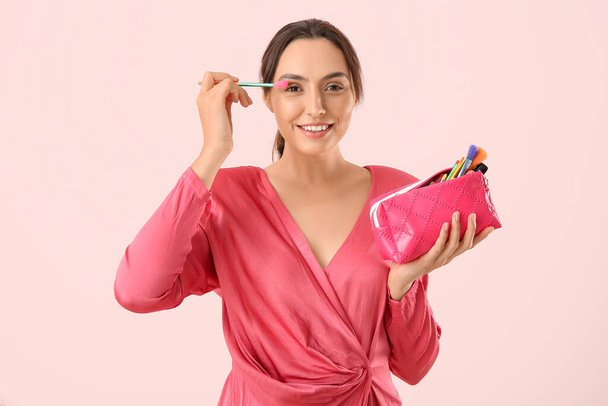 Krásná mladá žena s kosmetickou tašku plnou make-up produktů a kartáč na růžovém pozadí - Fotografie, Obrázek