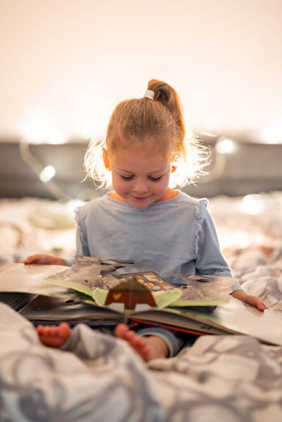 Маленькая девочка читает всплывающие книги в домашней постели в рождественской обстановке с огнями фона. Высокое качество фото - Фото, изображение