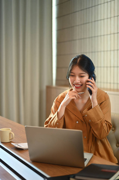 Happy Asian businesswoman having phone conversation and using laptop at modern office. - Photo, Image