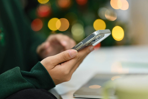 Femme dactylographier à la main sur smartphone sur fond de bel arbre de Noël avec des lumières en soirée. - Photo, image