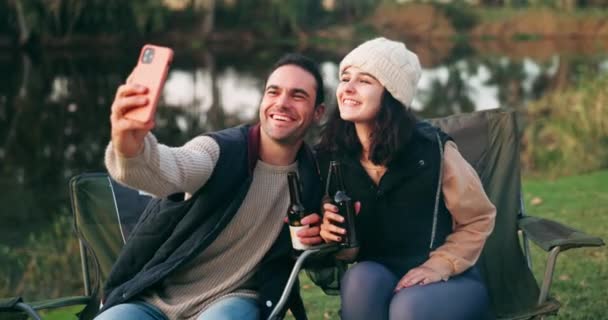 Selfie, κάμπινγκ και ζευγάρι με μπύρα δίπλα σε μια λίμνη για μια γιορτή και να ενημερώσετε τα μέσα κοινωνικής δικτύωσης των διακοπών μαζί. Διαδίκτυο, φωτιά και οι άνθρωποι ευτυχείς για μια online εικόνα στο υπαίθριο δάσος. - Πλάνα, βίντεο