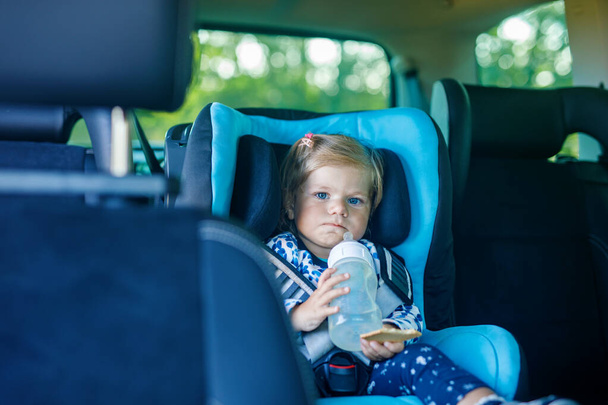 Adorable baby girl with blue eyes sitting in car safety seat. Toddler child going on family vacations and jorney. Smiling happy child during traffic jam, drinking milk from bottle and eating bisquit. - Photo, Image