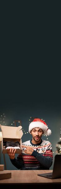 Video di unboxing. Uomo arabo in un cappello di Babbo Natale a casa mostra ai suoi abbonati nuovi occhiali VR durante una trasmissione in diretta. Influencer concetto di pubblicità. vista webcam. Ricevuto. Bandiera verticale - Foto, immagini