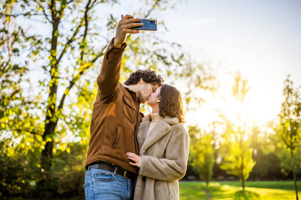 Retrato de casal amoroso feliz no parque ao pôr do sol. Eles estão beijando e tirando selfie. - Foto, Imagem