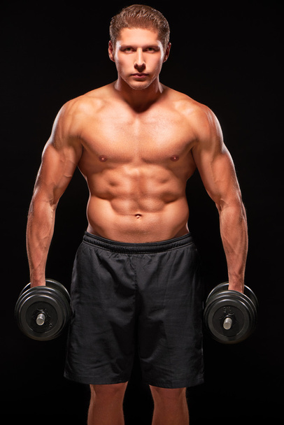 Serious muscular sportsman standing with dumbbells on black background - 写真・画像