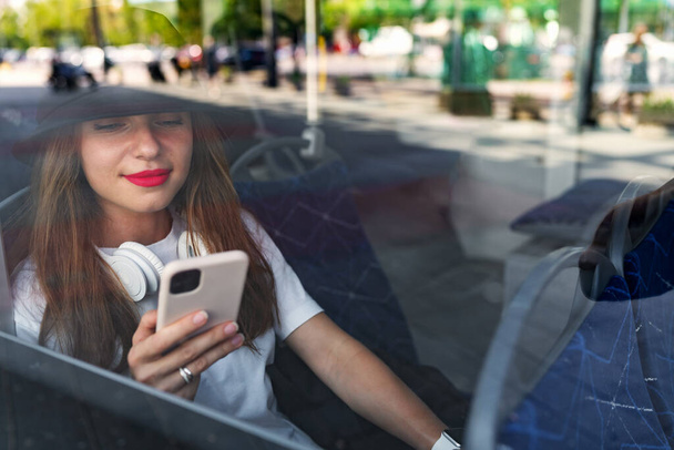 Kuva bussi-ikkunan lasin läpi, matkustaja nainen käyttää älypuhelinta istuessaan bussikuljetuksessa. - Valokuva, kuva