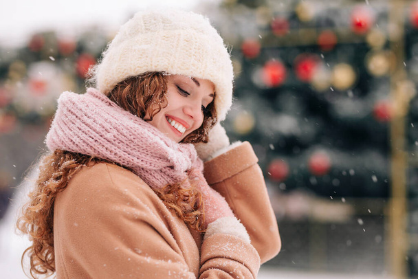 Sněžná městská krajina s mladou ženou v zimním oblečení, pózující před vánočním stromem. Zimní scéna s padajícím sněhem - Fotografie, Obrázek